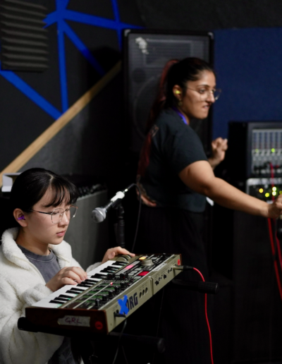 Two young people, one is playing a keyboard and the other is looking at an amp with wires and lights coming out of it.