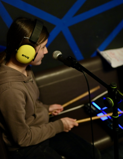 A young person wearing headphones, sat at a drum pad, holding two drumsticks.