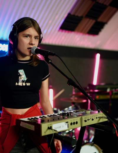 A young person wearing headphones standing at a keyboard and microphone. Their mouth is open as if they are singing into the microphone.