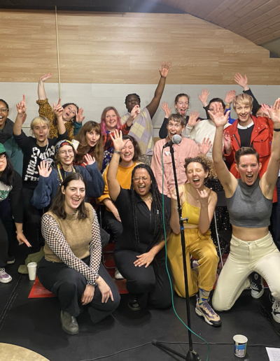 A group of diverse people of all ages with their hands up, all smiling and looking excited at the camera.